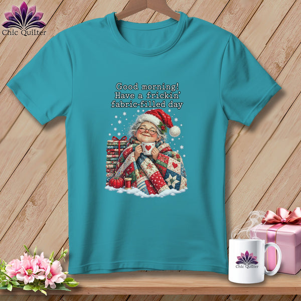 A teal t-shirt featuring an illustration of an elderly woman in a Santa hat, holding a mug and wrapped in a quilt.
