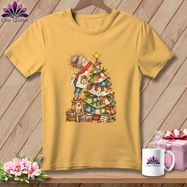 A mustard yellow t-shirt features a whimsical illustration of a woman placing a star on a quilted Christmas tree.