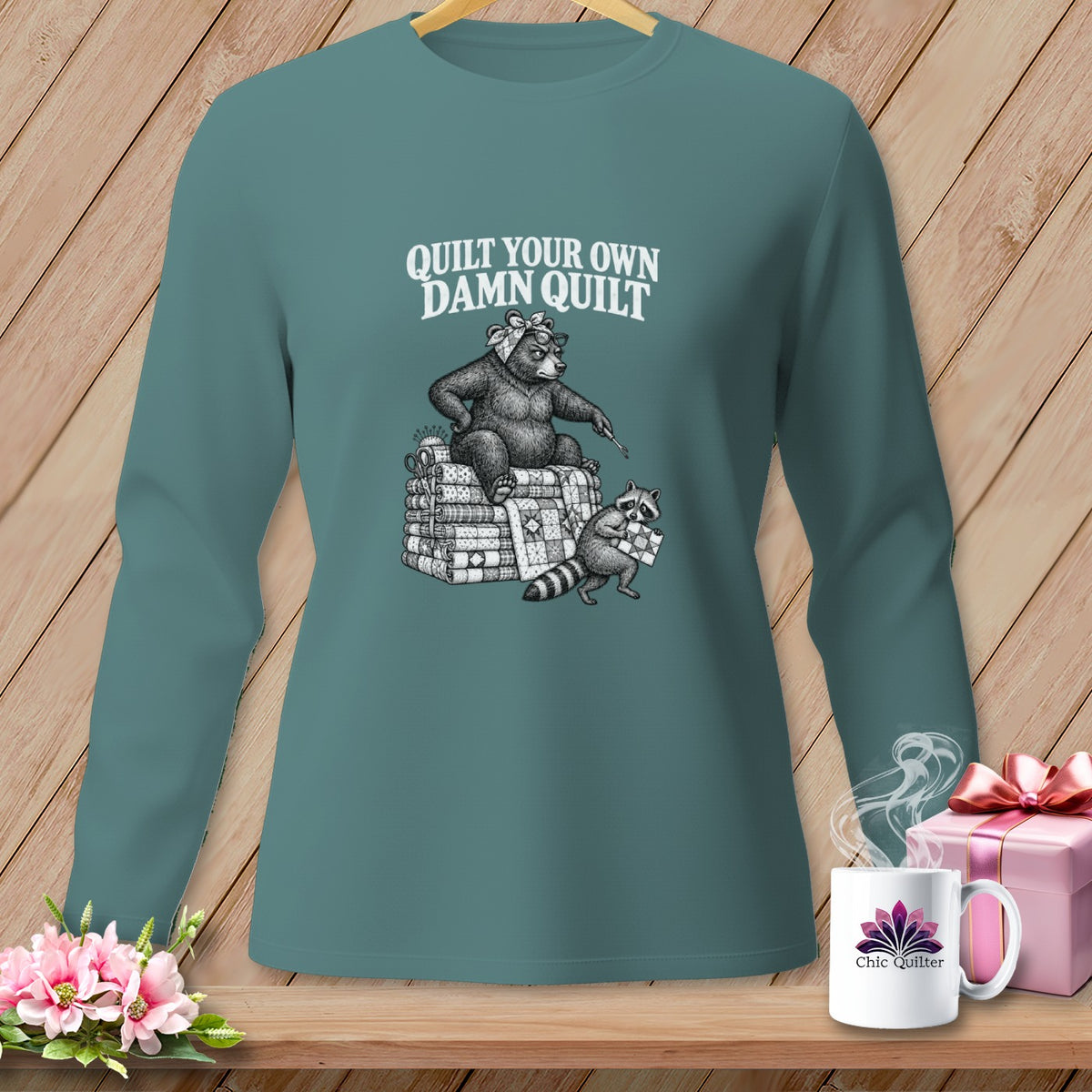 Teal long-sleeve t-shirt with a vintage illustration of a bear and raccoon quilting and the text Quilt Your Own Damn Quilt.