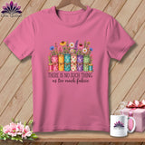 Pink short-sleeve t-shirt featuring rolled fabric graphic with flowers and text, hanging against wood background.