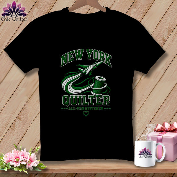 A black t-shirt hanging on a wooden background, featuring green text reading "New York Quilter" with an airplane and spool of thread graphic.
