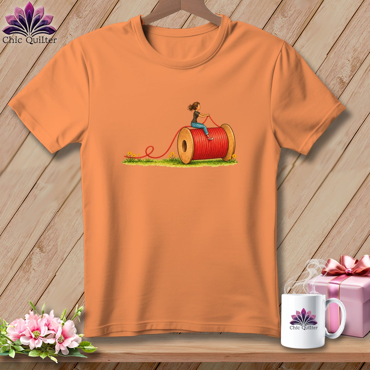 An orange t-shirt featuring a graphic of a person riding a giant spool of red thread.