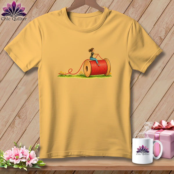 Yellow t-shirt featuring an illustration of a woman riding a large red spool of thread.