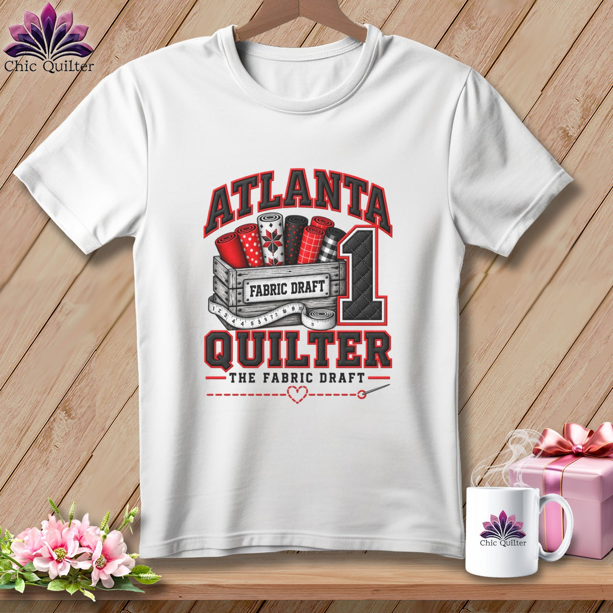 A white t-shirt hanging on a wooden background features a red and black design with fabric rolls and text reading Atlanta Quilter.