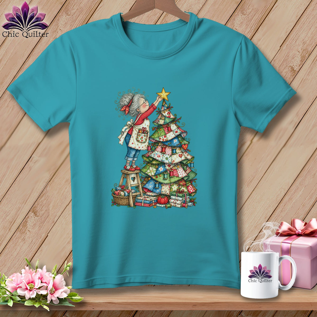 A teal short-sleeved t-shirt features a whimsical illustration of an elderly woman placing a star on a patchwork Christmas tree.