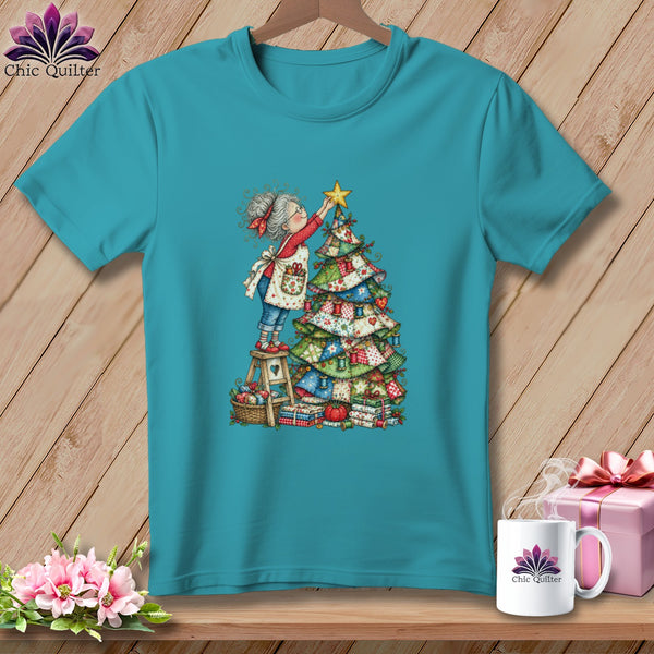 A teal short-sleeved t-shirt features a whimsical illustration of an elderly woman placing a star on a patchwork Christmas tree.