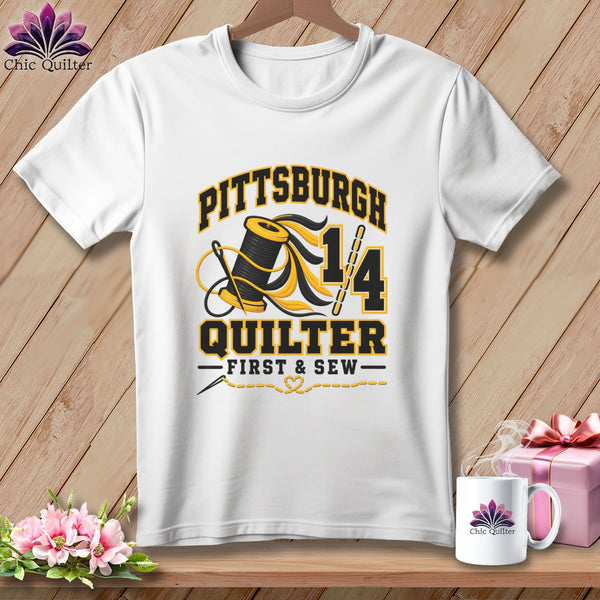 A white t-shirt hanging on a wooden background, featuring yellow and black Pittsburgh Quilter graphics with a spool of thread.