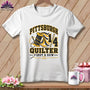 A white t-shirt hanging on a wooden background, featuring yellow and black Pittsburgh Quilter graphics with a spool of thread.
