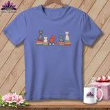 Periwinkle t-shirt featuring an illustration of five cats sitting on patterned cushions against a wooden background.