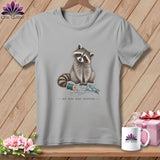 Grey t-shirt with a raccoon sitting by sewing supplies and the text 
