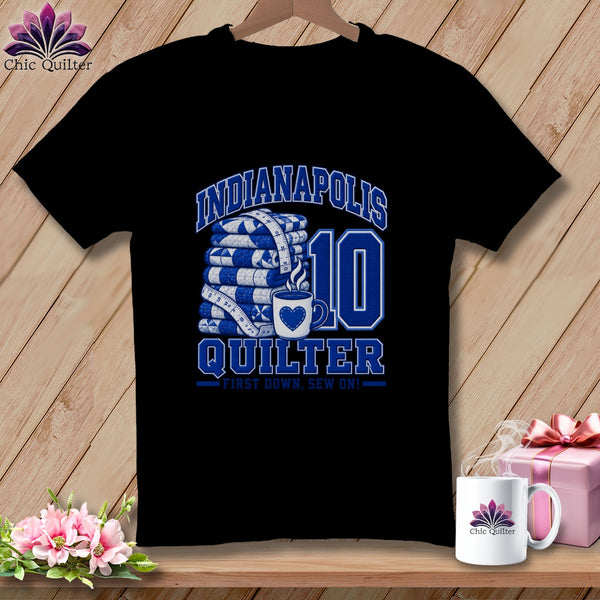 A black t-shirt featuring blue text reading Indianapolis Quilter First Down, Sew On with folded quilts, a measuring tape, and a coffee mug graphic.