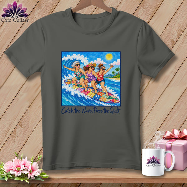 A charcoal grey t-shirt featuring a graphic of three older women surfing together on a colorful patterned surfboard with the text Catch the Wave, Piece the Quilt.