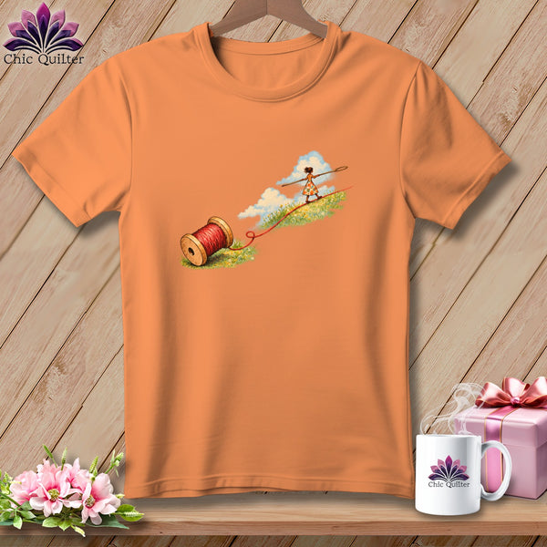 An orange crewneck t-shirt featuring an illustration of a spool of red thread and a tightrope walker.