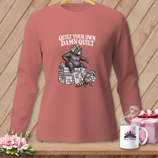Dusty rose long-sleeve shirt featuring an illustration of a bear and raccoon on stacked quilts with text that reads, Quilt Your Own Damn Quilt.