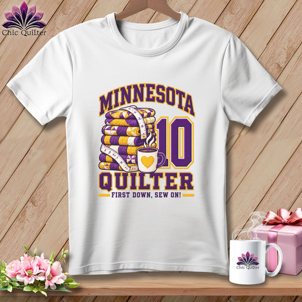 White t-shirt featuring a purple and yellow graphic with a stack of quilts, coffee mug, and text.
