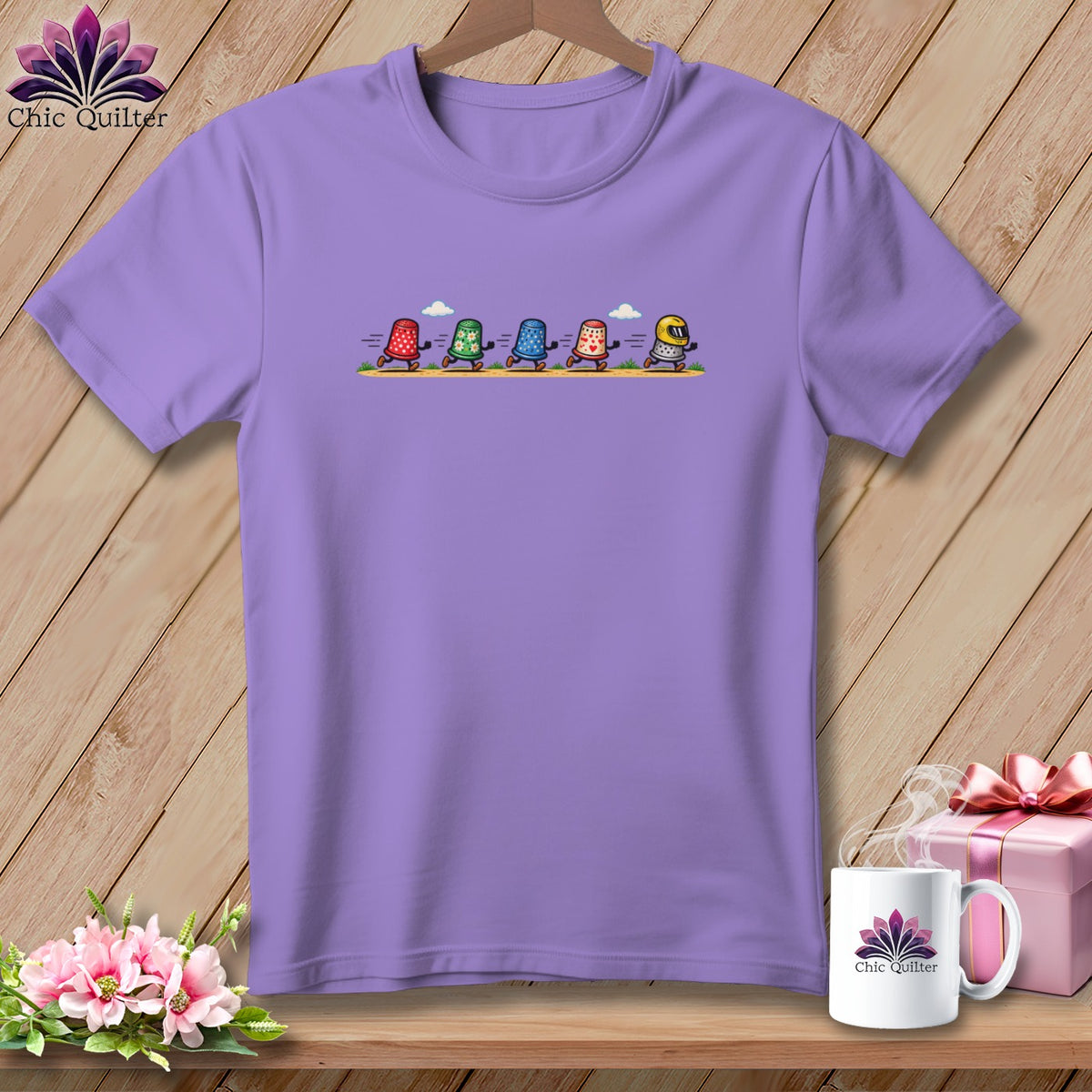 Light purple t-shirt featuring a graphic of five animated thimbles racing, displayed on a wooden background.