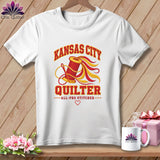 A white t-shirt hanging on a wooden background features a Kansas City Quilter graphic with a spool of thread.