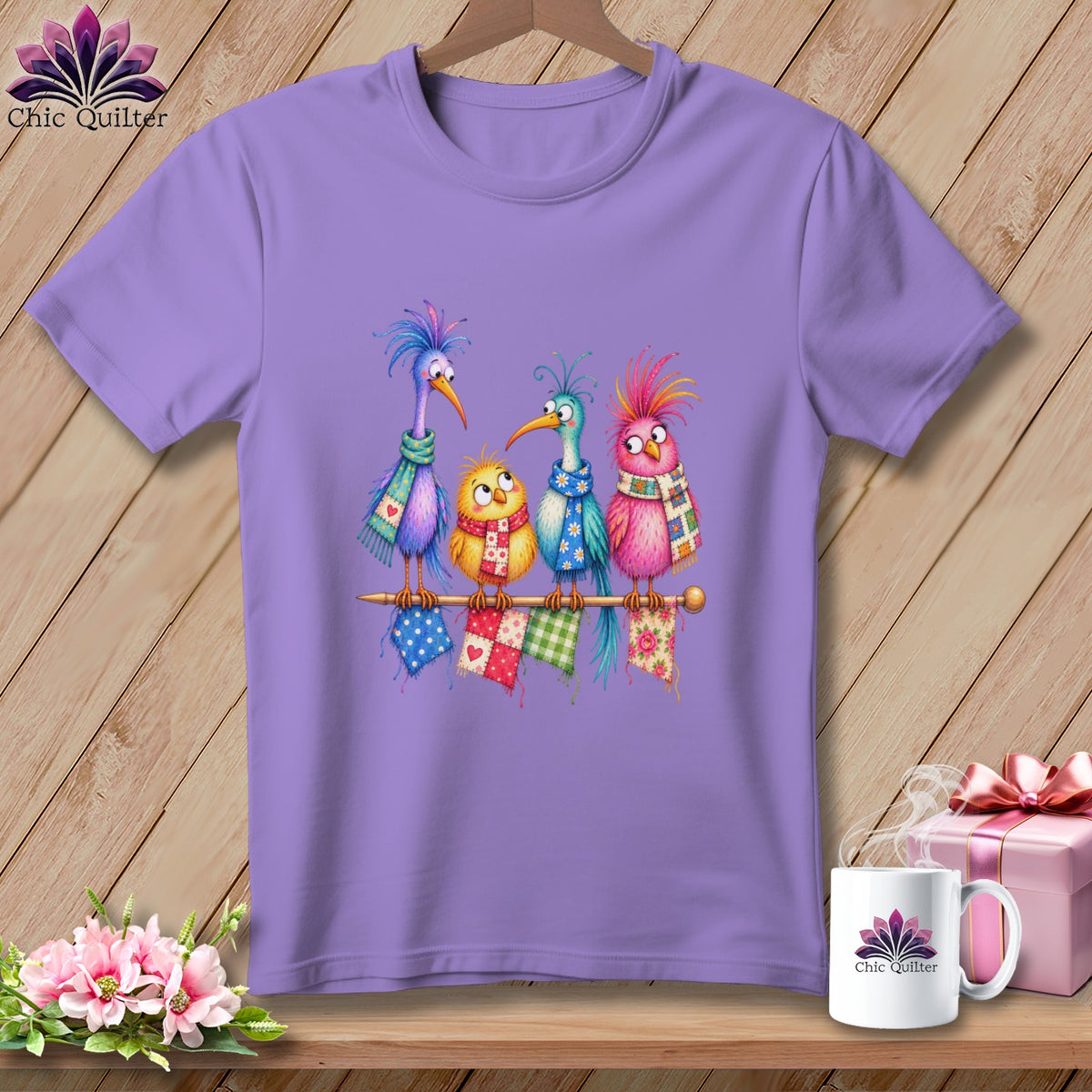 A light purple t-shirt featuring a colorful illustration of four quirky birds sitting on a knitting needle.