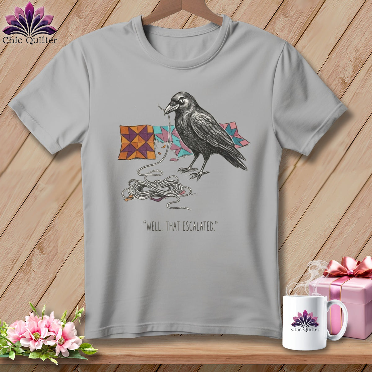 Grey t-shirt with a drawing of a crow holding thread and colorful quilt blocks, with text.