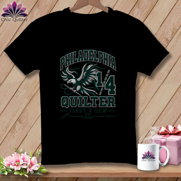 A black t-shirt featuring a green and white graphic with an eagle holding a needle, text reading Philadelphia Quilter 1/4, and a sewing theme.