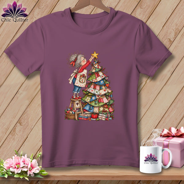 A purple t-shirt featuring a cartoon illustration of an elderly woman placing a star on a quilt Christmas tree.