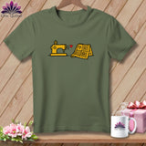 An olive green t-shirt with a cartoon graphic of a yellow sewing machine and quilt connected by a red heart.