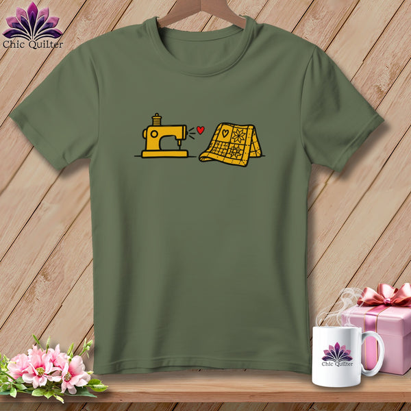 An olive green t-shirt with a cartoon graphic of a yellow sewing machine and quilt connected by a red heart.