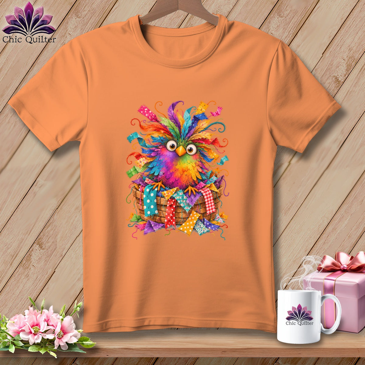 An orange t-shirt displays a graphic of a colorful, feathered bird nestled inside a basket of fabric scraps.