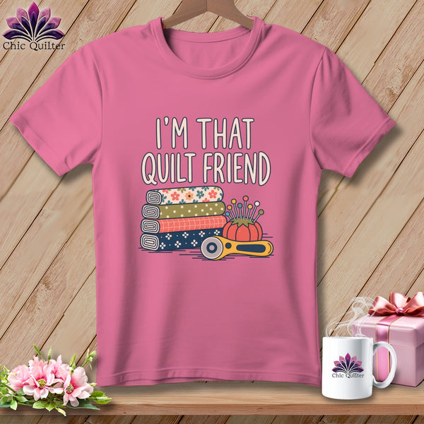 Pink t-shirt with the text I'M THAT QUILT FRIEND above stacked fabric rolls, a pincushion, and a rotary cutter.