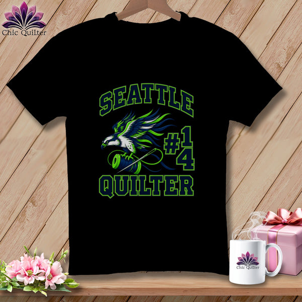 A black t-shirt features a green and blue graphic of an eagle, a spool of thread, and the text Seattle Quilter #14.
