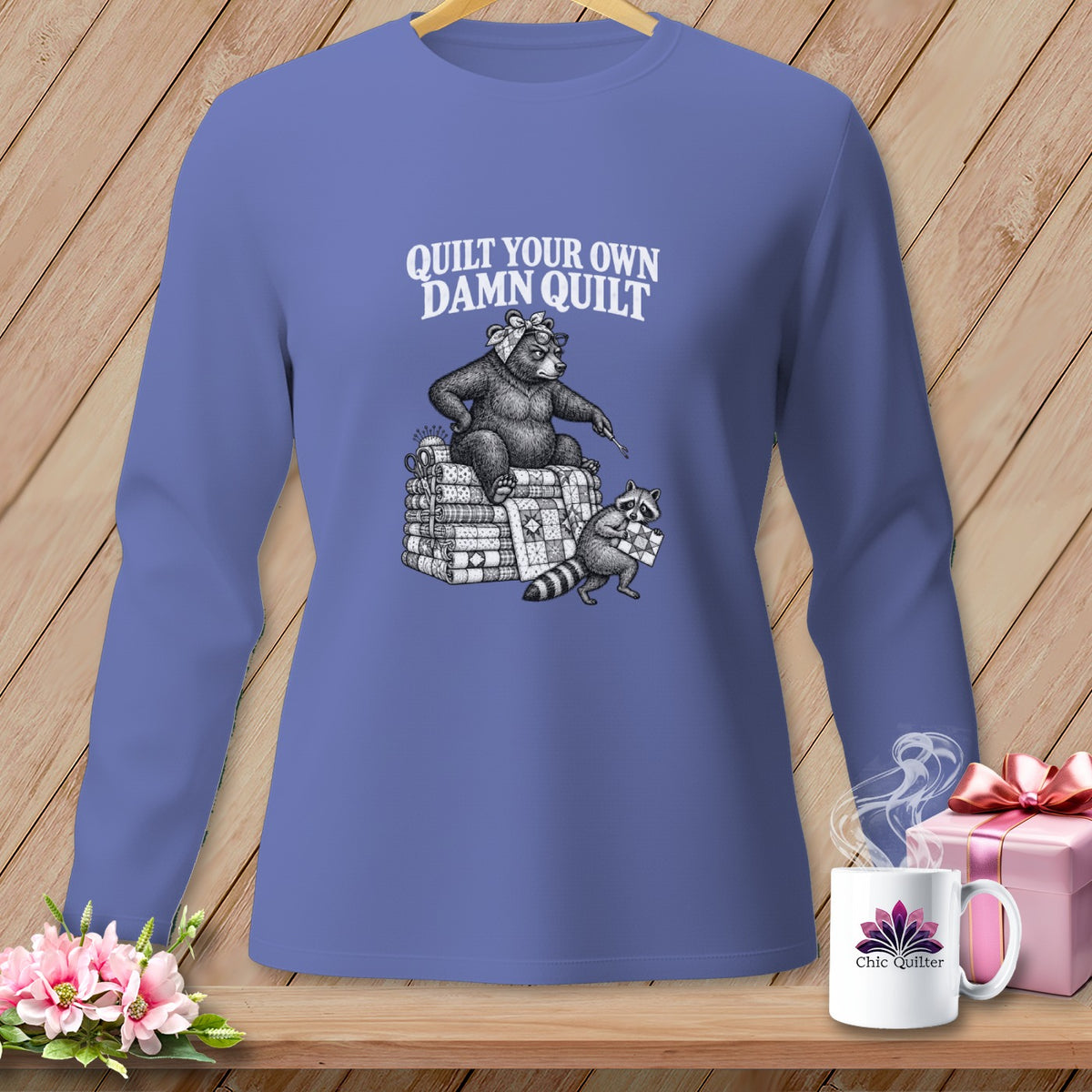Periwinkle long-sleeve shirt with a vintage graphic of a bear and raccoon quilting and the text Quilt Your Own Damn Quilt.