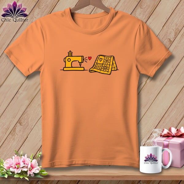 An orange short-sleeved t-shirt with a graphic of a yellow sewing machine and folded quilt connected by a red heart.
