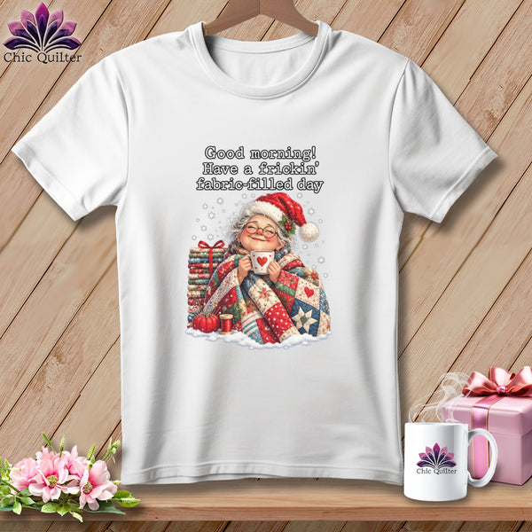 White t-shirt with a graphic of an elderly woman in a Santa hat holding a mug, wrapped in a quilt, and text reading, "Good morning! Have a frickin' fabric-filled day."