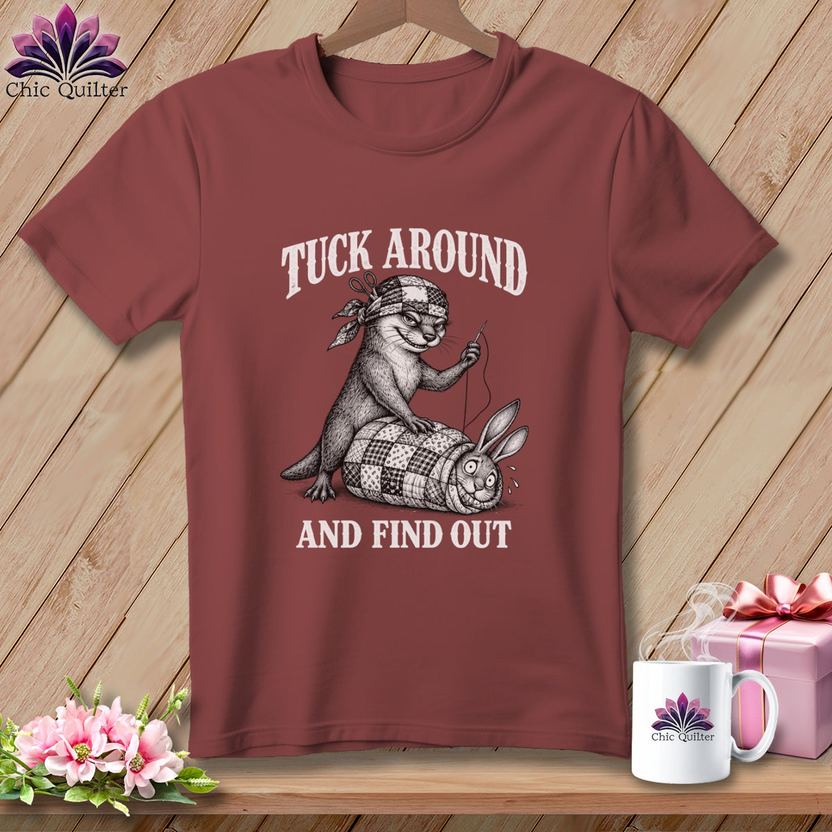 Maroon t-shirt featuring a graphic of an otter sewing a rabbit into a quilt, with text reading TUCK AROUND AND FIND OUT.