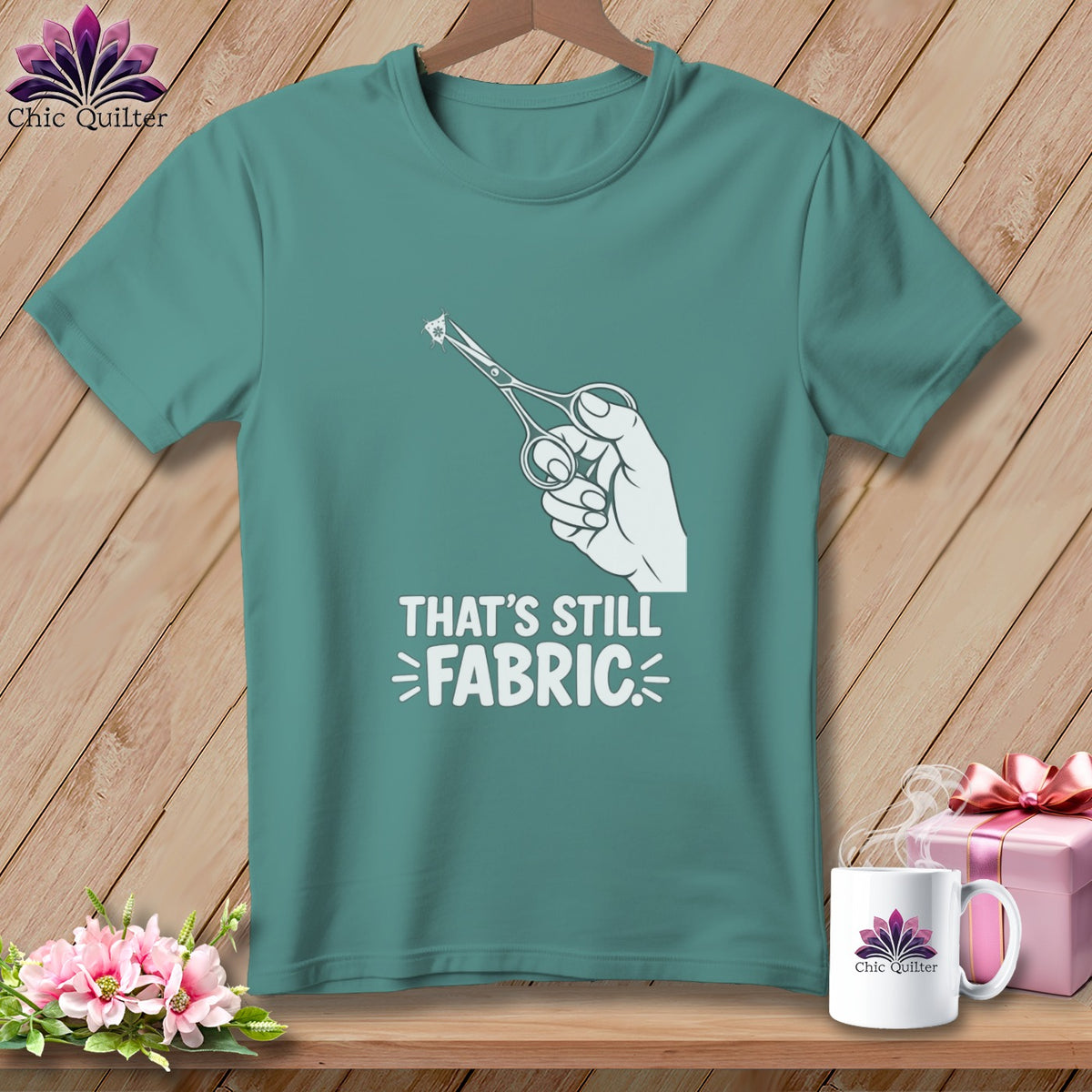 Teal t-shirt with white graphic of hand holding scissors cutting fabric and text "THAT'S STILL FABRIC".