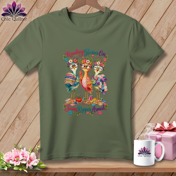 An olive green t-shirt displays an illustration of three colorful birds wearing glasses and flower crowns.