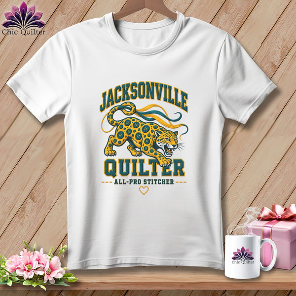 A white t-shirt hanging on a wooden background, featuring a Jacksonville Quilter graphic with a jaguar illustration.