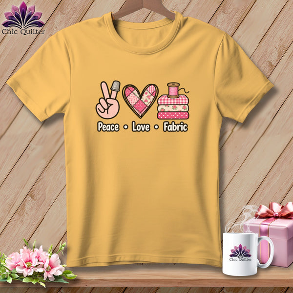 A gold t-shirt hanging on a wooden background, featuring the text Peace Love Fabric with sewing-themed graphics.