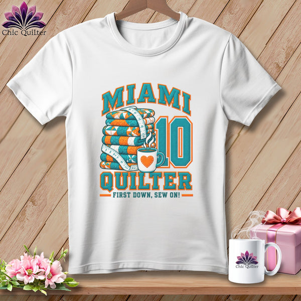 A white t-shirt hanging on a wooden wall features a graphic with stacked quilts, a coffee mug, and the text Miami Quilter First Down, Sew On.