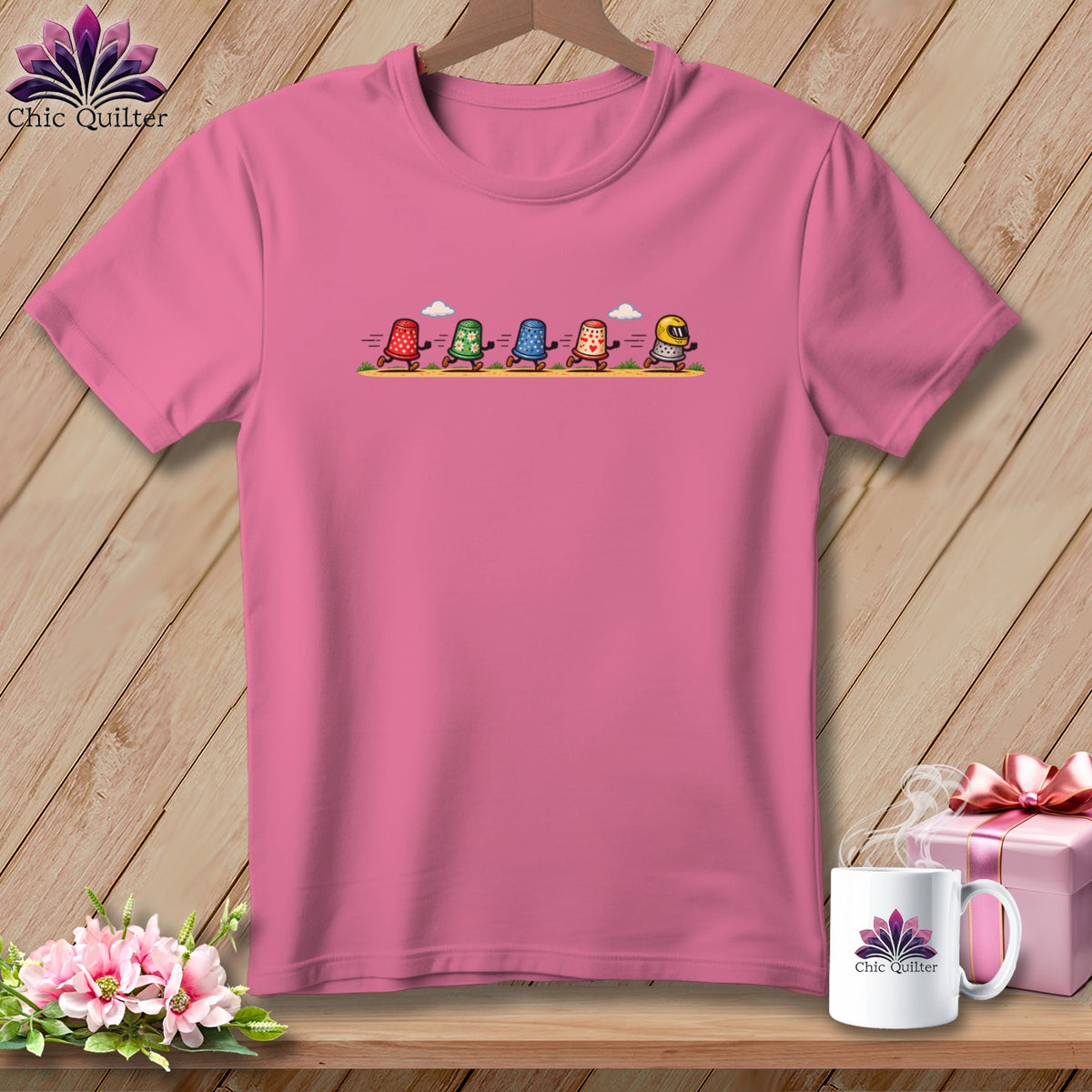 Pink crew neck t-shirt featuring a graphic of five running thimbles on a wooden background.