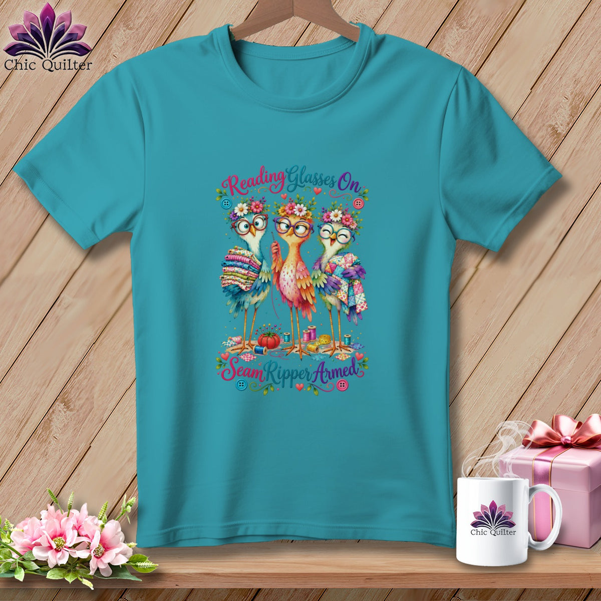 Teal t-shirt featuring a cartoon design of three birds wearing glasses and floral crowns holding sewing supplies.