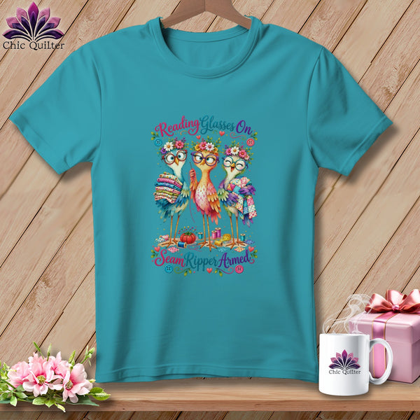 Teal t-shirt featuring a cartoon design of three birds wearing glasses and floral crowns holding sewing supplies.