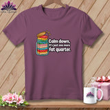 Plum-colored t-shirt on a wooden background, featuring a graphic of a jar filled with folded fabrics and text reading, Calm down, it's just one more fat quarter.