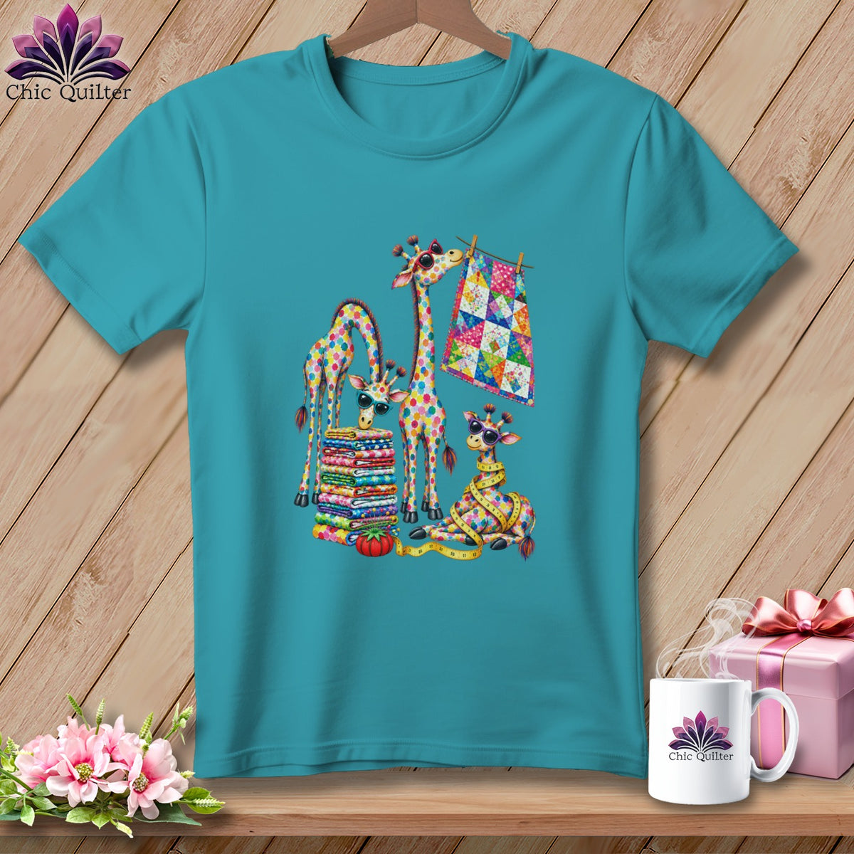 Teal crewneck t-shirt featuring a colorful graphic of giraffes sewing and stacking vibrant quilts.