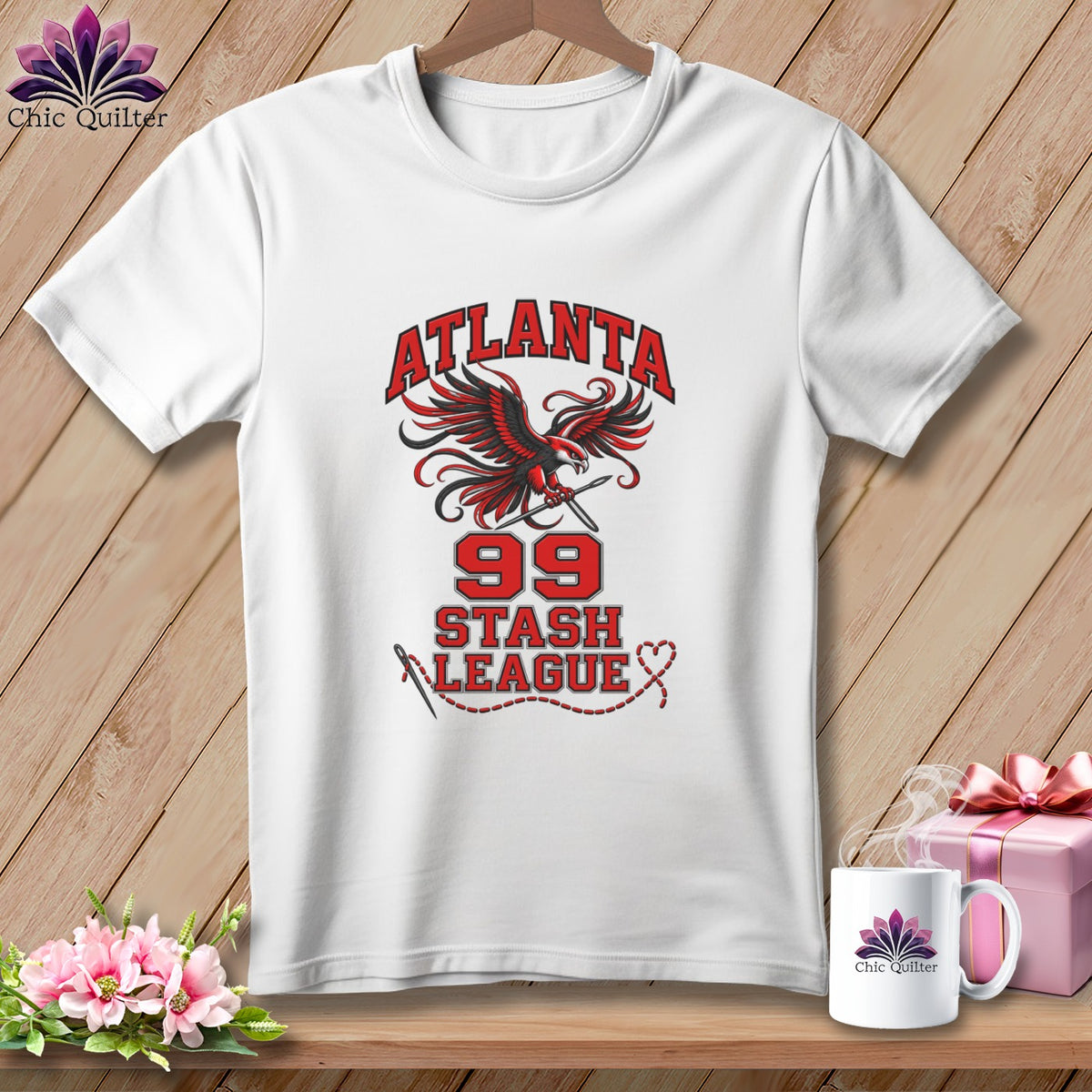 A white t-shirt hanging on a wooden background features a graphic with a red and black falcon, the text "ATLANTA 99 STASH LEAGUE," and a needle and thread design.