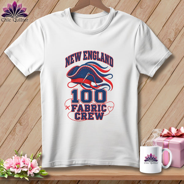 A white t-shirt hanging on a wooden background, featuring blue and red embroidery of a pirate hat with text reading New England 100 Fabric Crew.