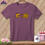 A dark purple short-sleeved t-shirt featuring a yellow sewing machine, a red heart, and a yellow folded quilt graphic on the chest.