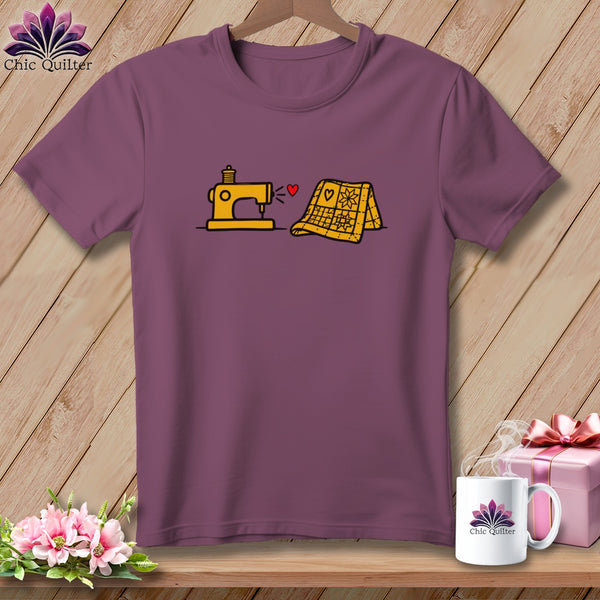 A dark purple short-sleeved t-shirt featuring a yellow sewing machine, a red heart, and a yellow folded quilt graphic on the chest.