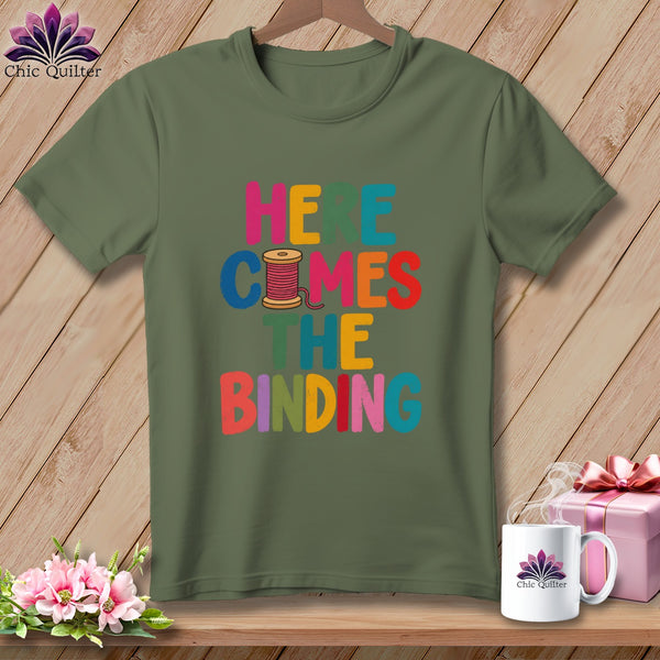Olive green crewneck t-shirt featuring the colorful text Here Comes the Binding and a spool of pink thread.