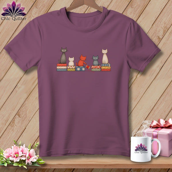 Purple short-sleeved t-shirt featuring an illustration of five cats sitting on patterned blocks of fabric.